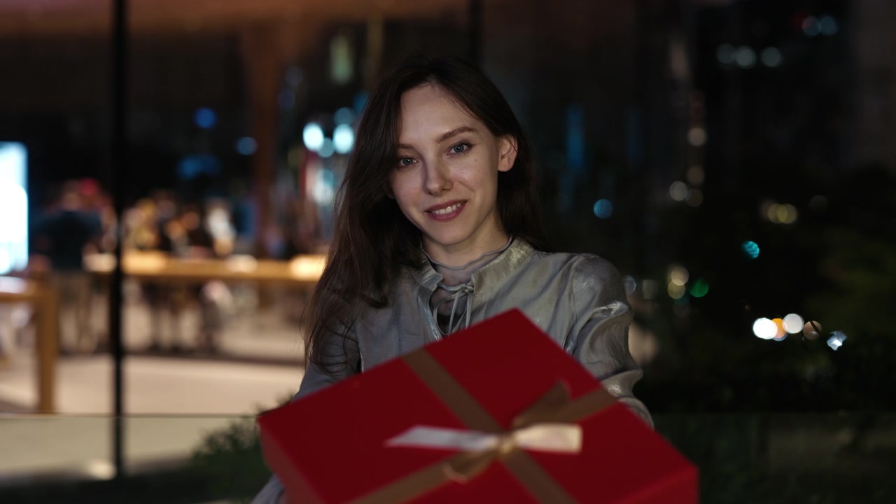 Giovane donna sorridente presenta una scatola regalo rossa al chiuso di notte
