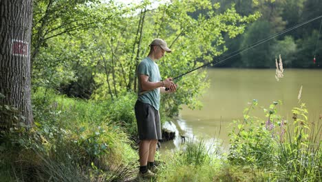 young angler at the edge of a murky lake surrounded by trees turns the winch and reels in fishing line