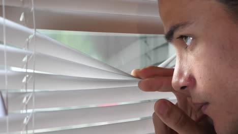 An-Attractive-South-American-Male-Adult-Inconspicuously-Peers-Through-Window-Blinds-Surveilling-the-Outdoors,-Static-Tripod-Close-Up