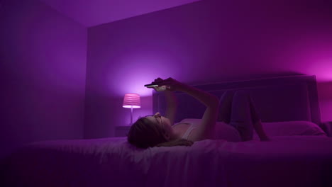 girl laying on back browses social media on smart phone late at night in bedroom.