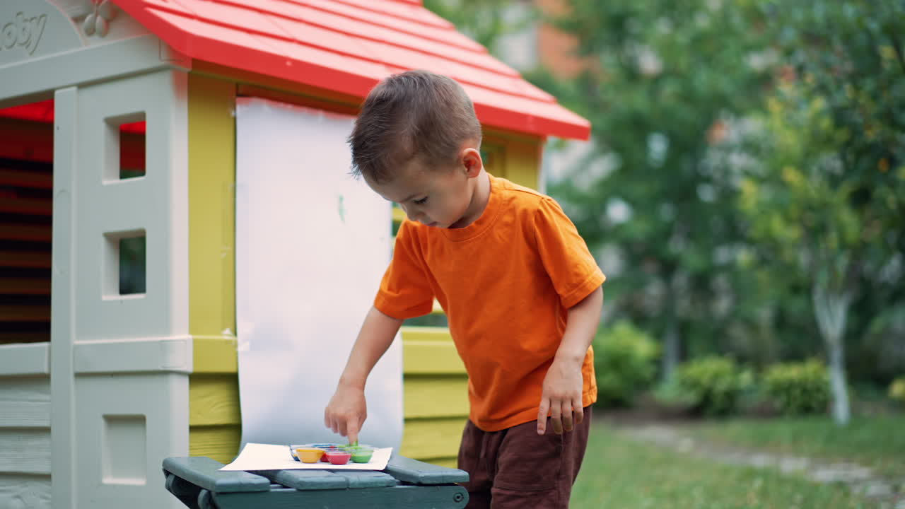 Little Caucasian boy stands at the playhouse outdoors. Kid paints on the paper attached to the wall. Painting with finger using no brush. Low angle view.