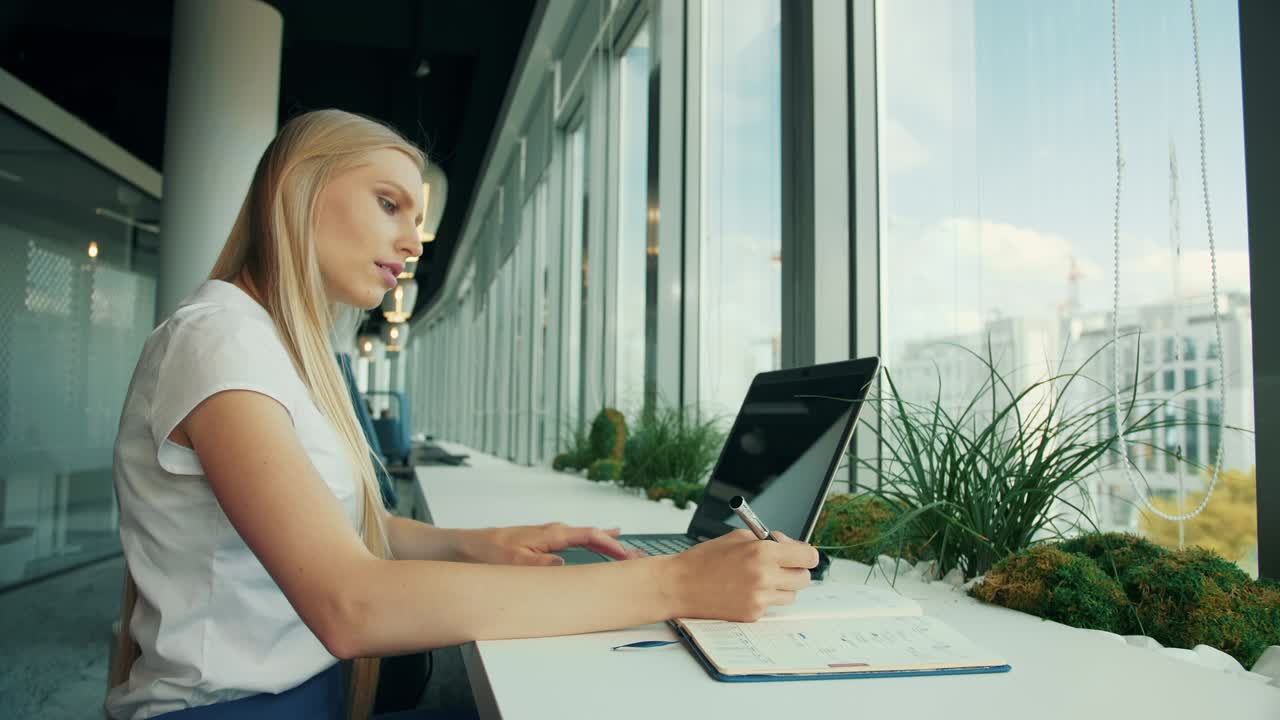 Donna in abito formale con laptop e documenti. Vista laterale di una donna d'affari che scrive su un foglio mentre è seduta con il laptop a un tavolo vicino alla finestra in un ufficio moderno