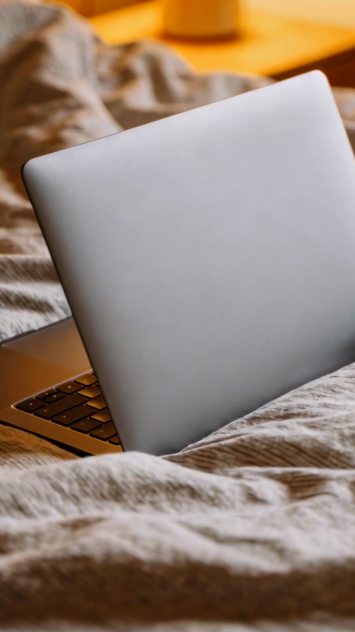 Silver laptop resting ajar on rumpled duvet, camera shifting and showing keyboard for tech promo