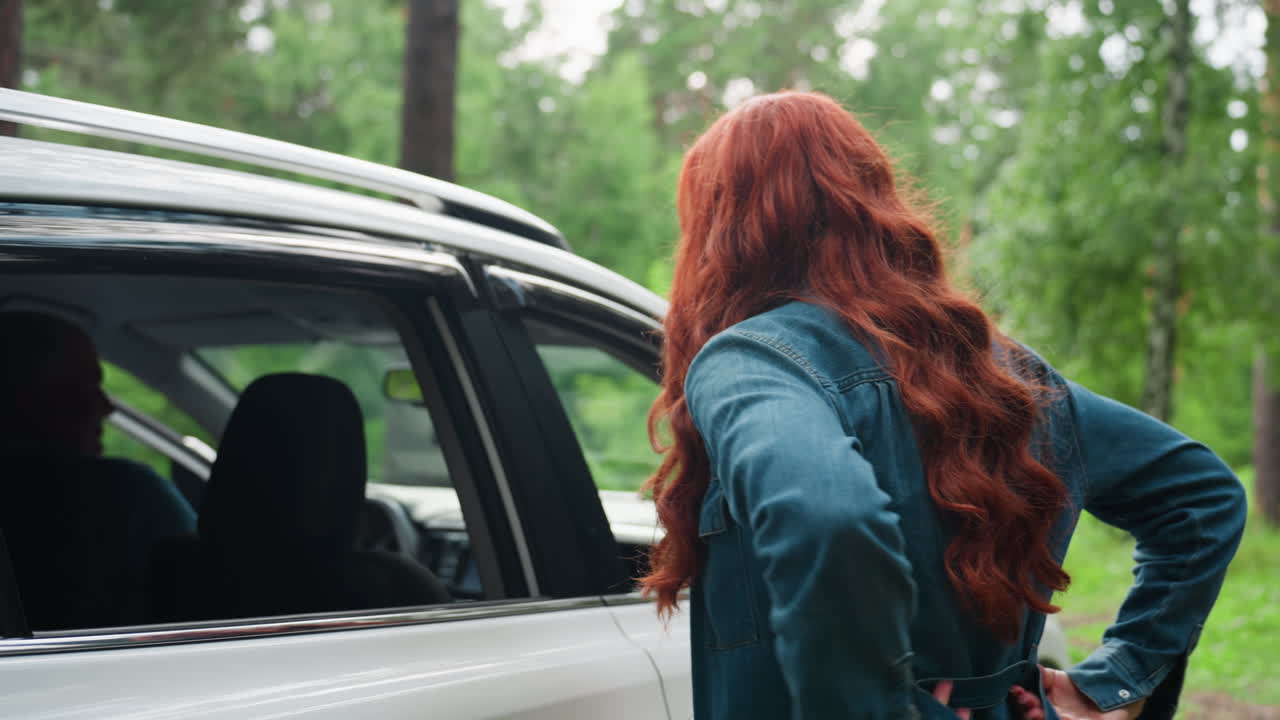 Stylish mother with long red hair stands beside white family SUV adjusting her dress while husband enters car from driver side, surrounded by lush green forest, calm outdoor family travel moment