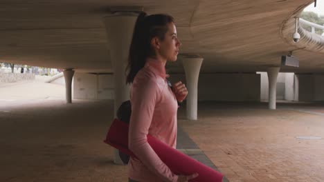 Caucasian-woman-walking-under-bridge-with-yoga-mat