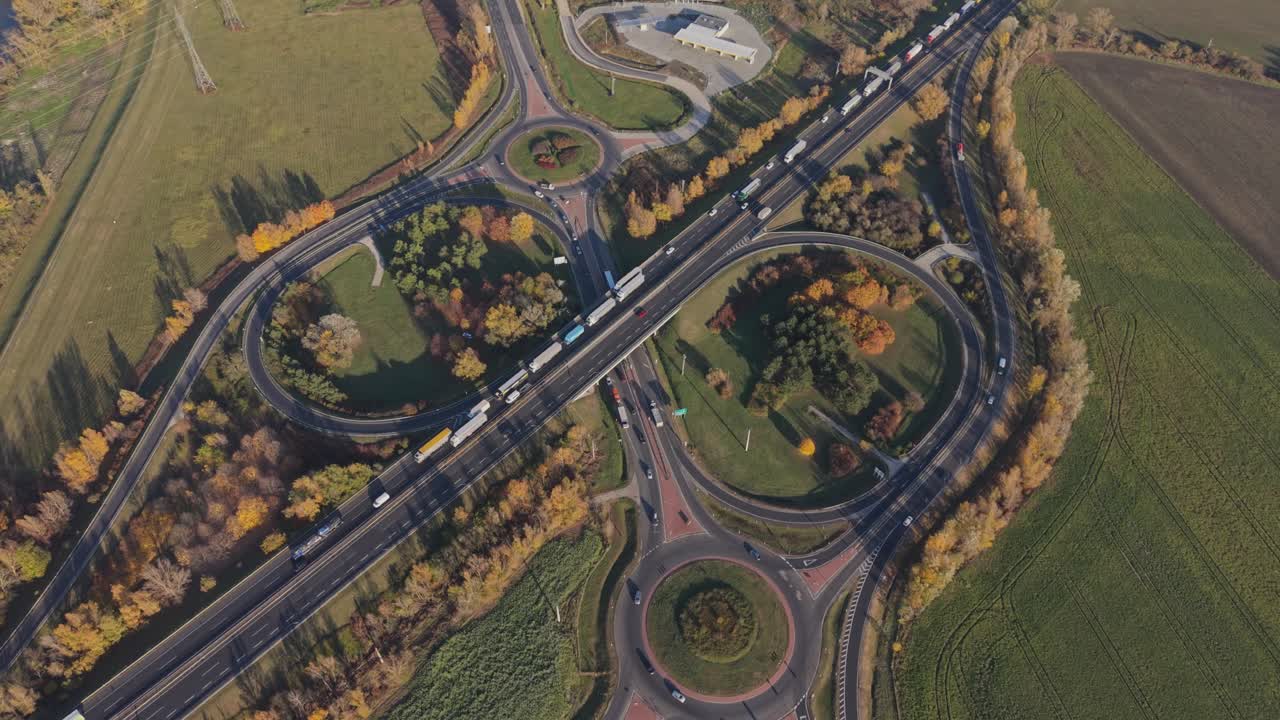 A high-angle drone footage of a large highway interchange with multiple roundabouts and overpasses. Trucks and cars travel through the junction surrounded by green fields and autumn-colored trees