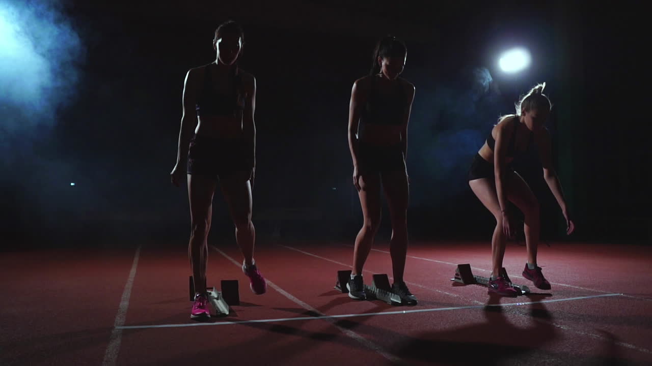 Female runners at athletics track crouching at the starting blocks before a race. In slow motion.