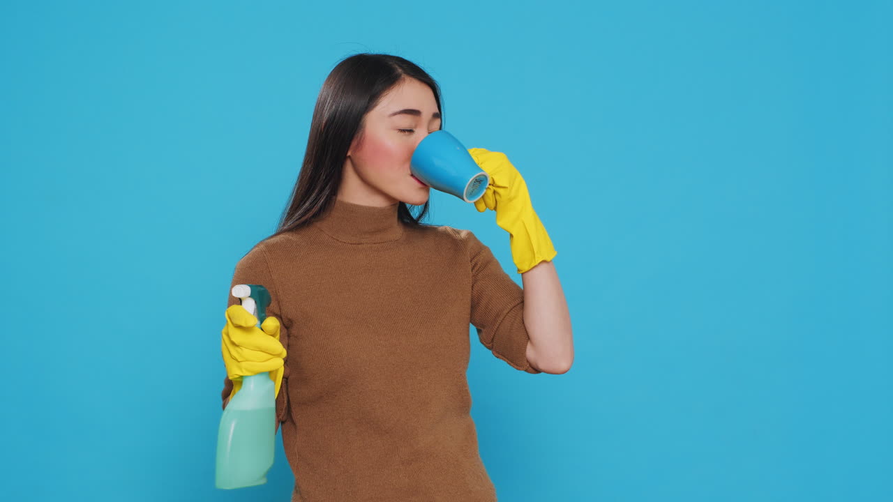 Happy housekepper enjoying cup of coffee while cleaning house with chemical spray bottle