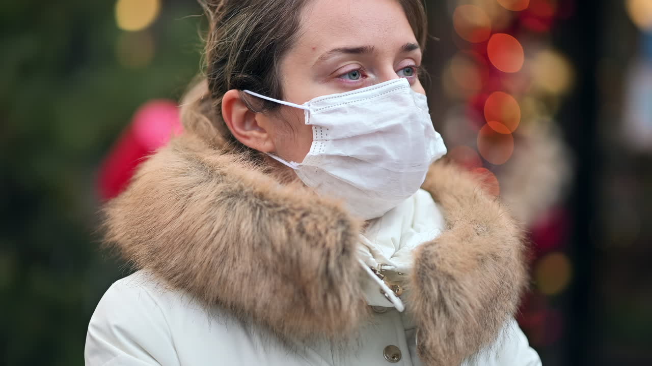 A young woman stands outside, wearing a mask while watching the lively city around her. The cold weather brings a cozy atmosphere with festive decorations lighting up the streets