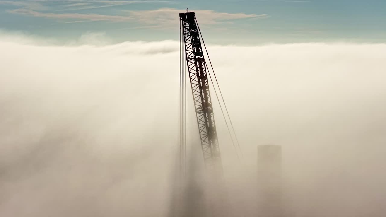 Aerial pull back view of a silhouetted construction crane building a wind turbine, emerging through dense clouds in a Latvian wind farm at sunset