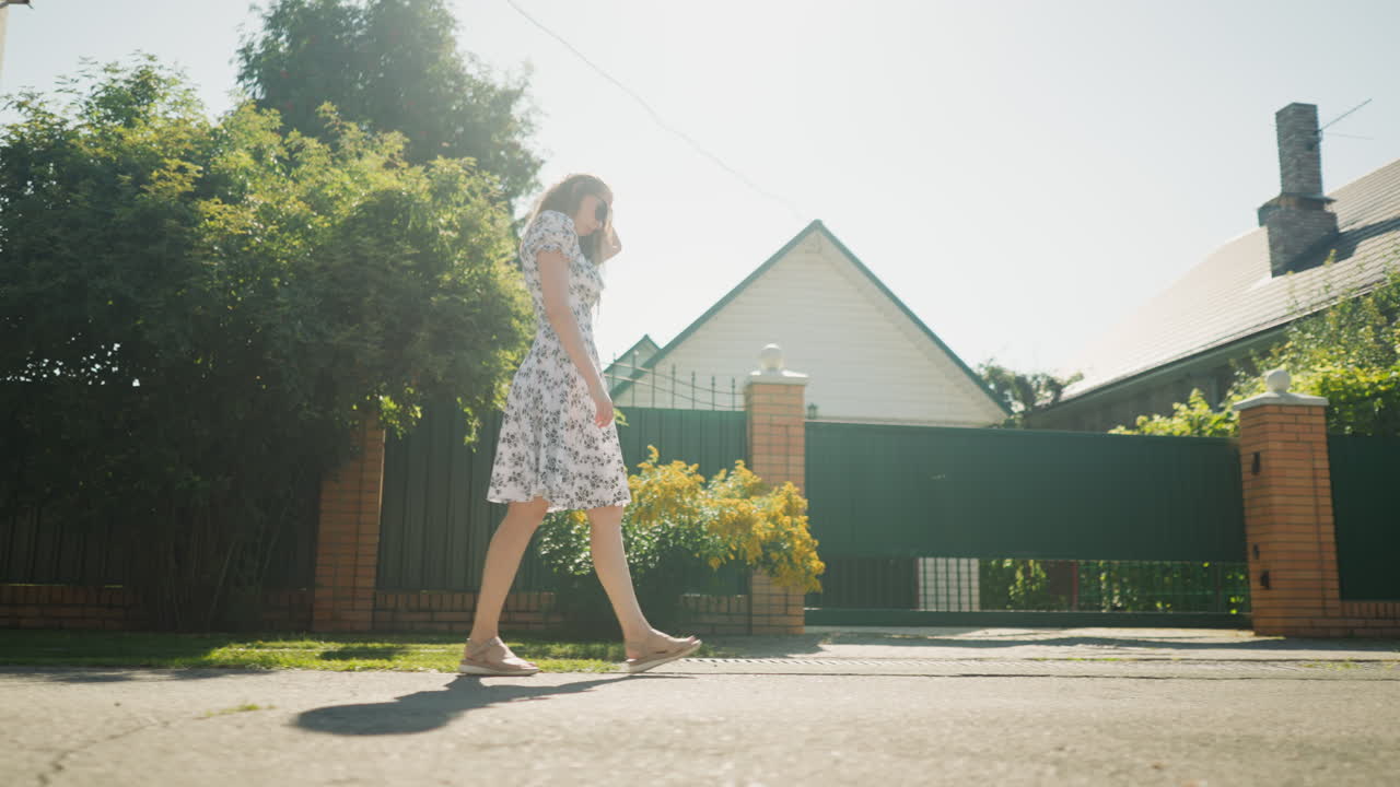 Young woman in floral dress walking calmly under intense midday sunlight, passing by green fence and utility pole, with shadows cast on road and suburban house in background