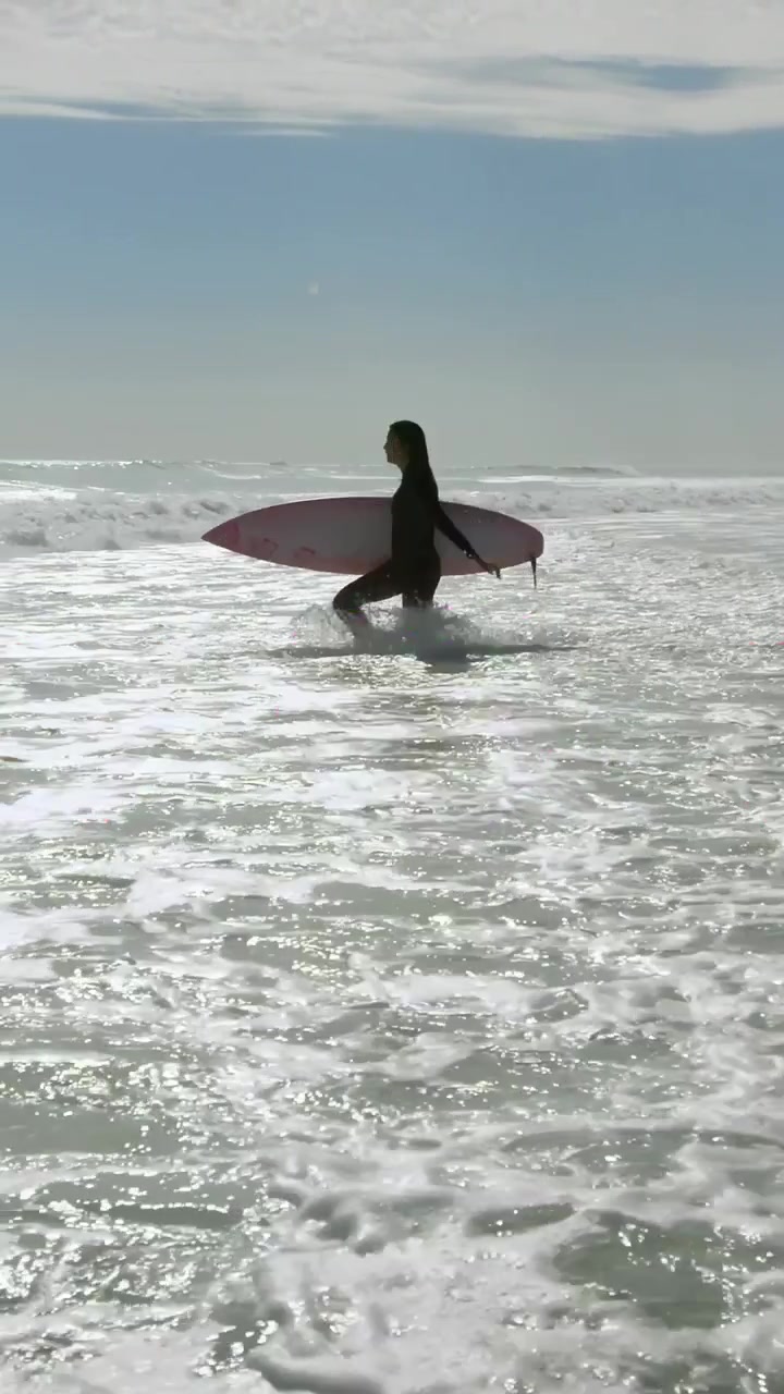 Video verticale: Donna che si addentra tra le onde tenendo una tavola, mostrando cuori con LOVE o FOREVER per annunci di surf