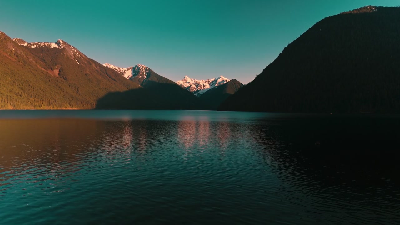 Vista dal basso sull'acqua calma e scura di un lago, che si increspa dolcemente riflettendo la brillante luce dorata del tramonto che si irradia da lontane cime montuose innevate, ripresa con drone aereo ad alta quota.