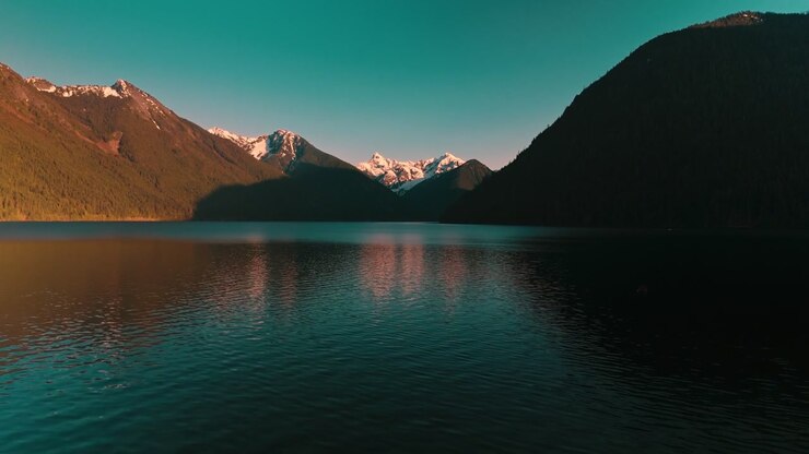 Vista dal basso sull'acqua calma e scura di un lago, che si increspa dolcemente riflettendo la brillante luce dorata del tramonto che si irradia da lontane cime montuose innevate, ripresa con drone aereo ad alta quota.