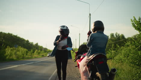 two women in helmets stand by the roadside, with one seated on a motorcycle while the other walks toward her, in the background, cars approach, and a biker waves as they pass