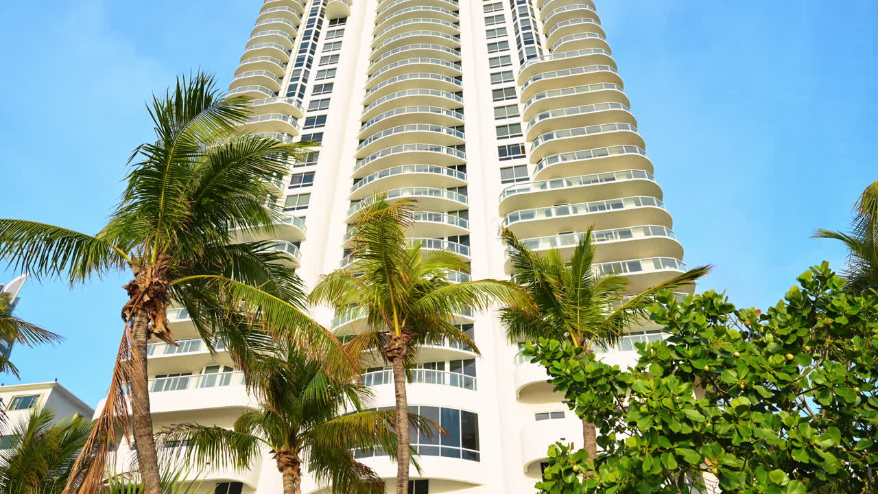 Varandas de um edifício residencial branco emolduradas por palmeiras tropicais. Vista em ângulo baixo de um edifício de apartamentos moderno e branco com varandas curvas atrás de palmeiras verdes em Miami.