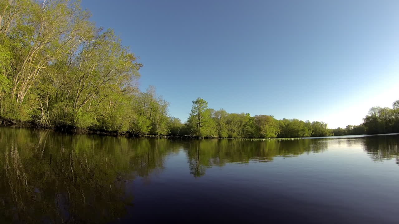 Boat-mounted forward trucking shot advances through glassy still water. Wooded shoreline is mirrored in water.