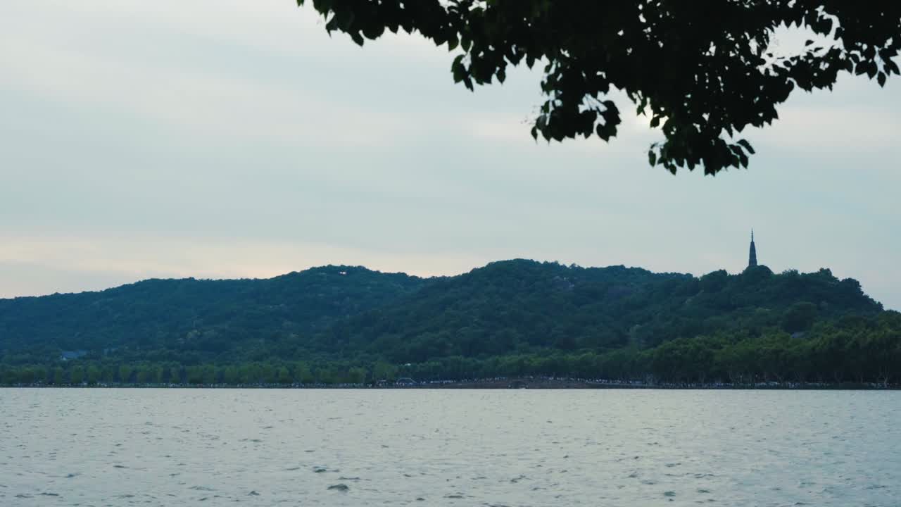 Serene aerial footage of Hangzhou's West Lake with Leifeng Pagoda in the distance. Captures tranquil waters, lush hills, and soft daylight, evoking a peaceful atmosphere.