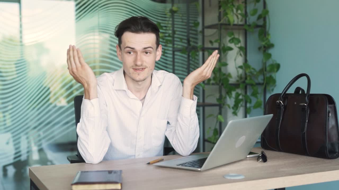멋지고, 젊고, 세련된 남자가 비즈니스 옷을 입고, 현대적인 사무실의 책상에 앉아, 비디오 통화를 통해 누군가와 이야기하고 논쟁하고 있습니다.