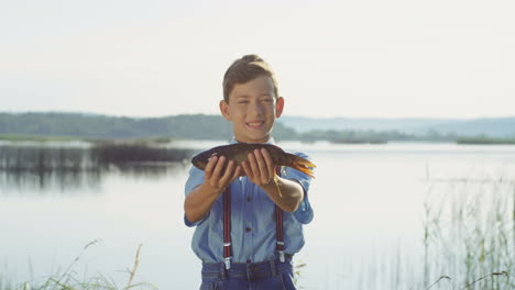 portrait of a cute teenage boy standing on the lake shore and holdinga fish while looking at the camera