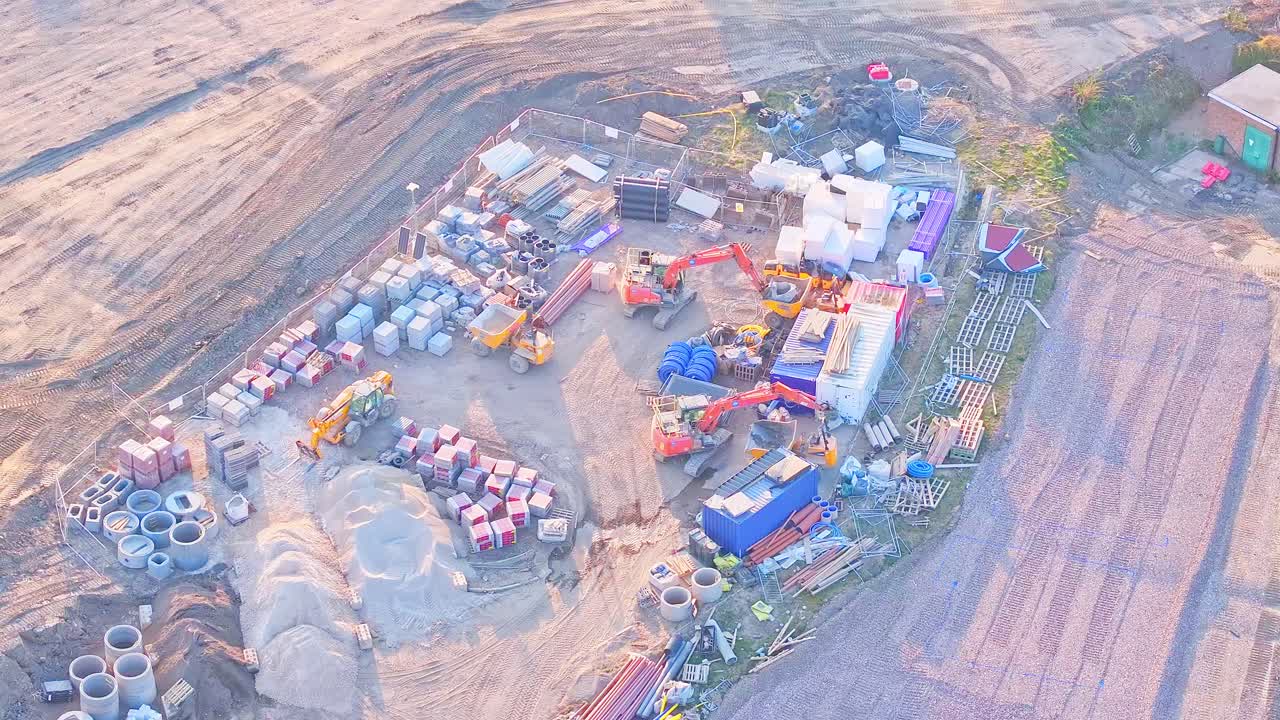 Busy construction material yard, showcasing machinery, building supplies, and equipment surrounded by bare earth in Wombwell, South Yorkshire, England, High-angle drone orbit