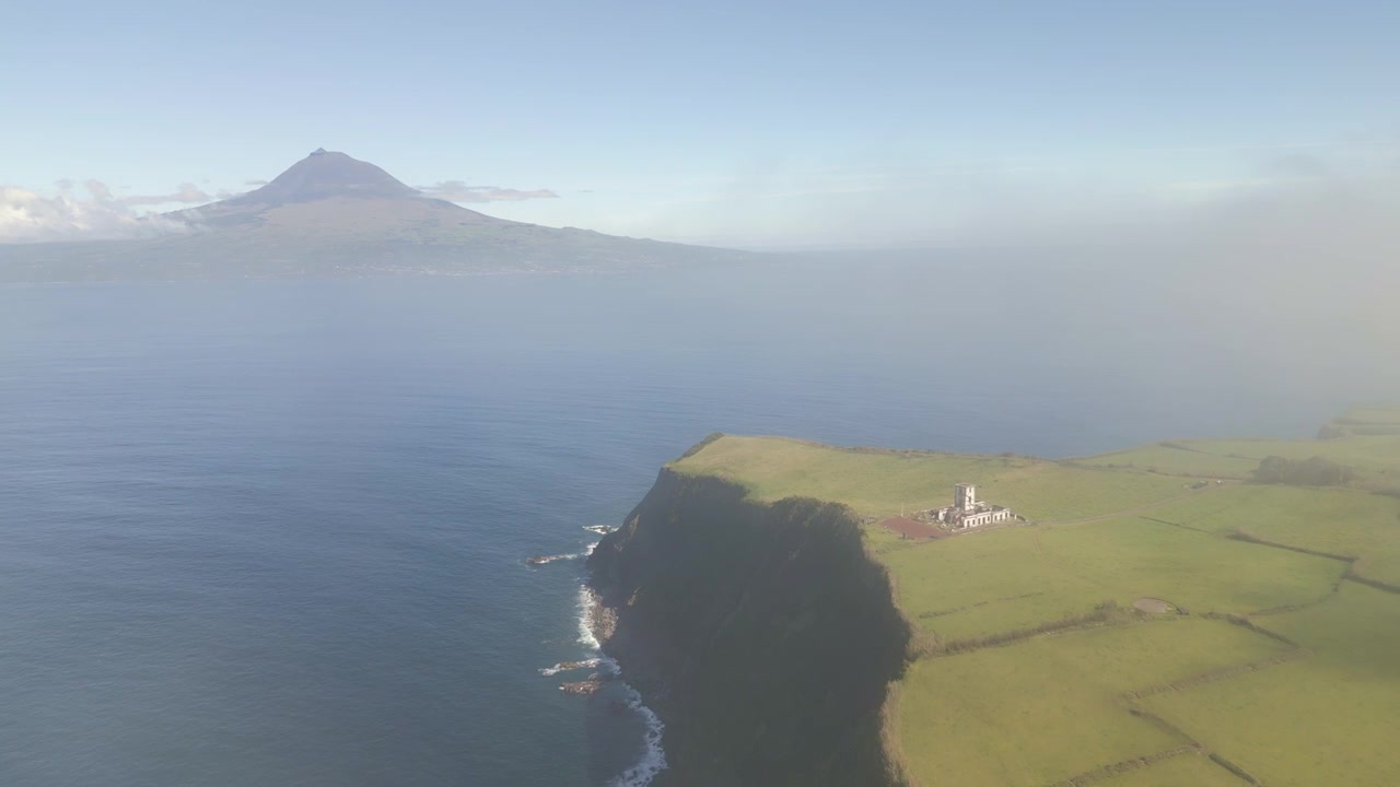Vista aerea, mattina nebbiosa del Faro di Ribeirinha a Faial, Azzorre, Portogallo, con il Monte Pico che si erge attraverso l'Atlantico e la foschia che si sposta sulla campagna costiera