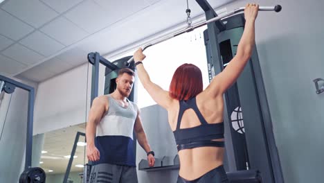 back view of strong woman exercising on a modern simulator. red-haired sportswoman pulling the bar down and her instructor controls the process in the gym.