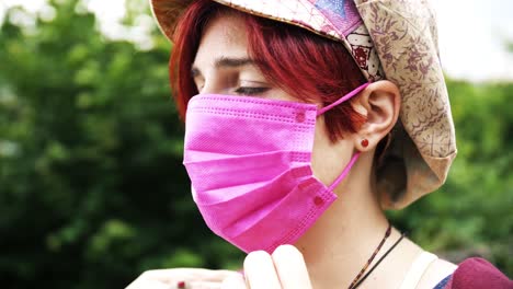 side view of young red haired girl with colorful hat putting on a medical pink face mask with green park in background