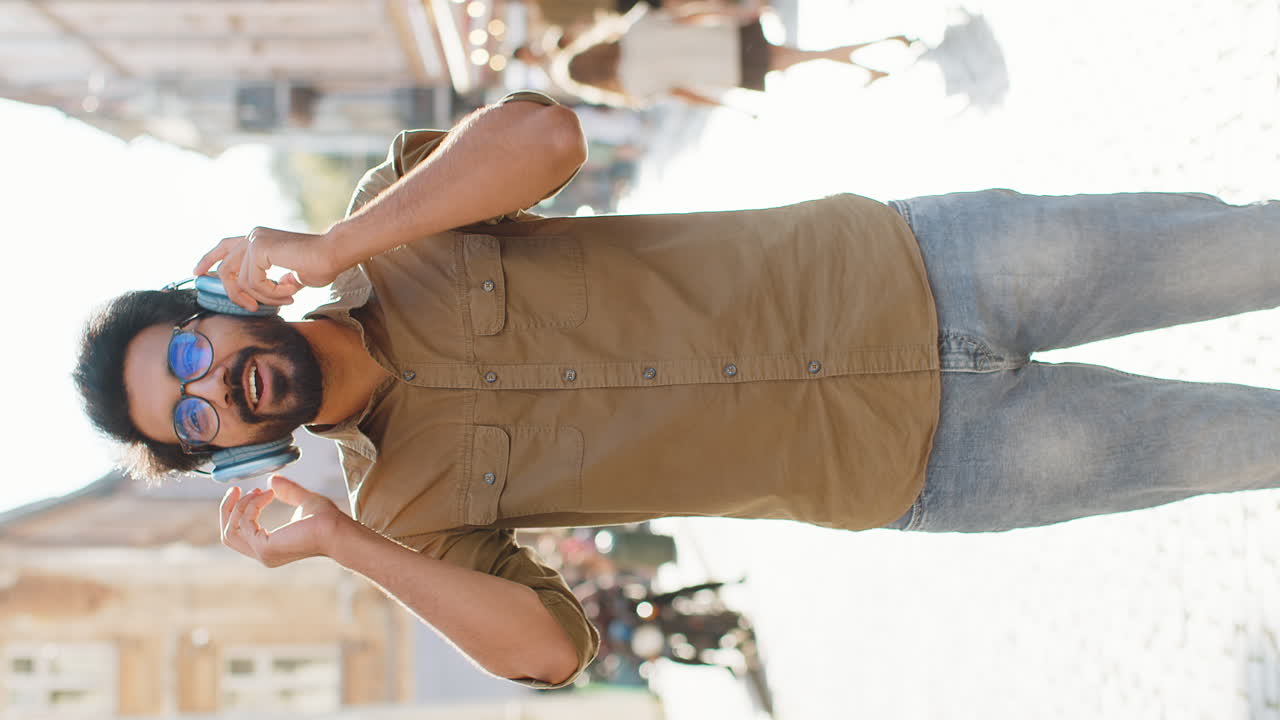 Happy young man in wireless headphones choosing listening music dancing outdoors city street