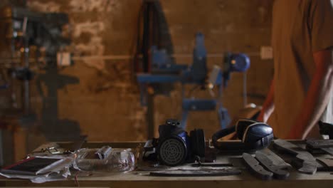 midsection of caucasian male blacksmith walking in workshop with tools on table