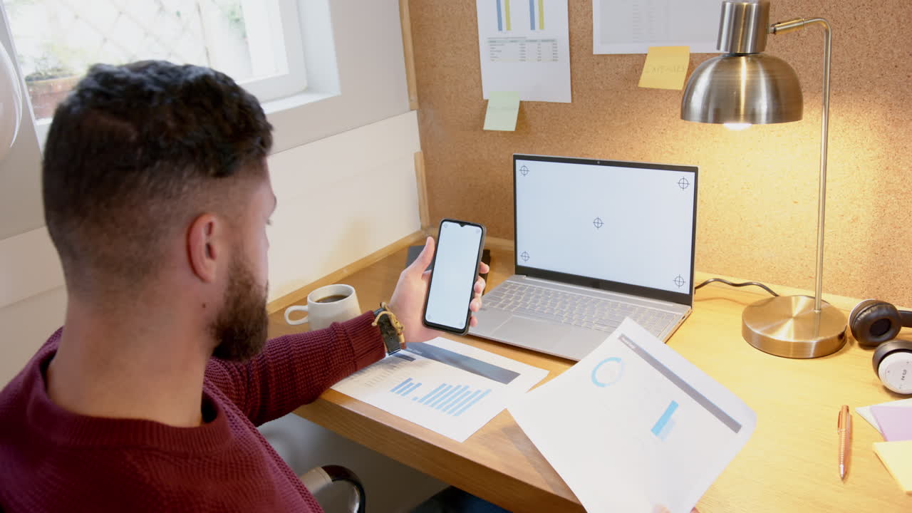 Holding smartphone and reviewing documents, man working from home office desk, copy space