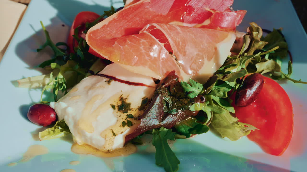 Close up of a gourmet salad with fresh mozzarella, cured meat slices, tomatoes, olives and mixed greens on a white plate in bright daylight