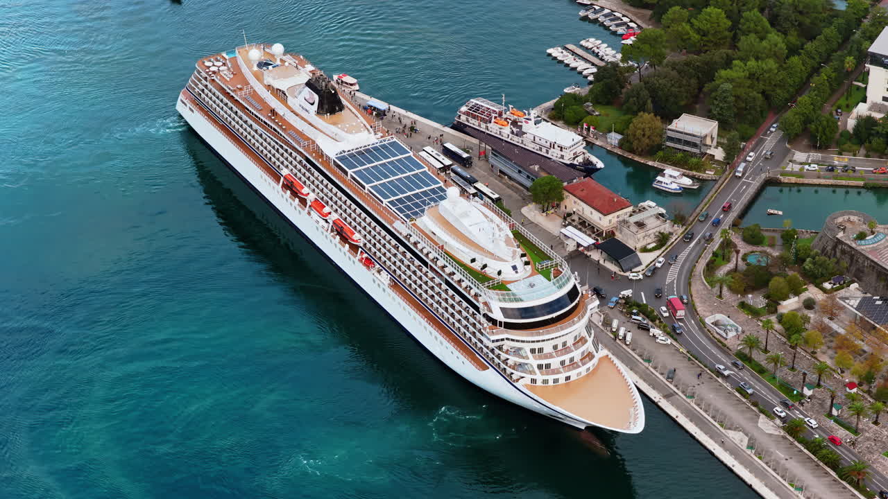 Aerial drone view of a large cruise ship docked at the Kotor port, with buses, marina, and waterfront infrastructure visible