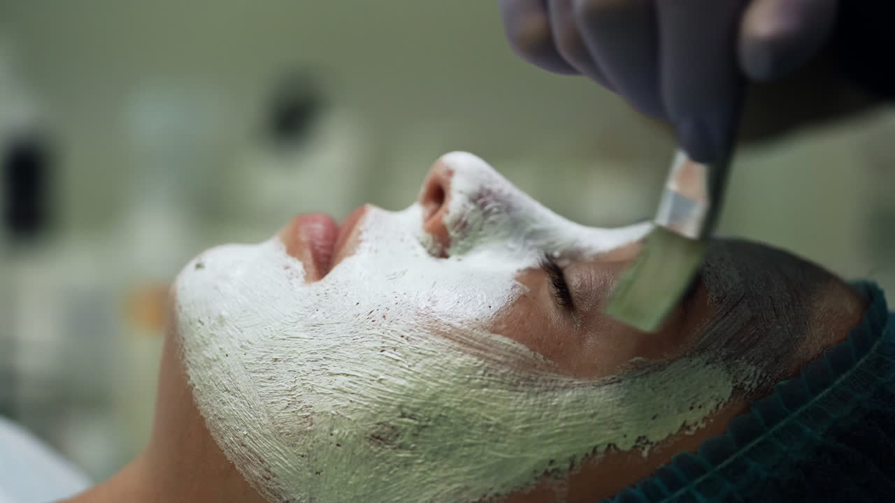 Beautician puts a white mask on the female client's face. Skin care and rejuvenation in modern beauty salon. Side view close up.