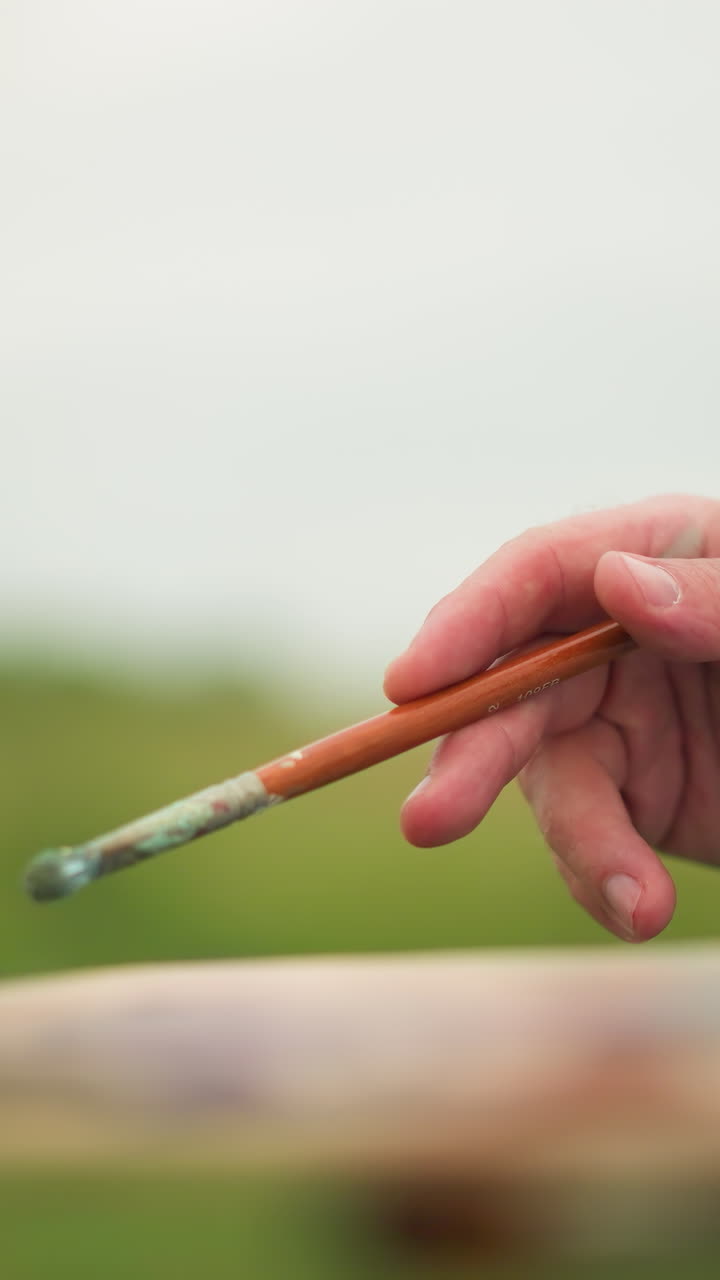 toma de primer plano de la mano de un artista sosteniendo un pincel con una paleta borrosa visible en el fondo. la imagen captura el delicado agarre y enfoque, en un fondo natural sereno y borroso