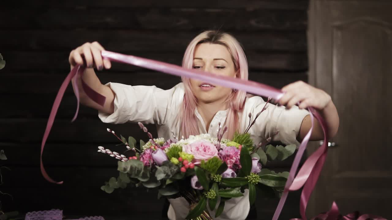 Professional girl florist binds the bottom of the bouquet with silk pink ribbons. Dark studio. Slow motion