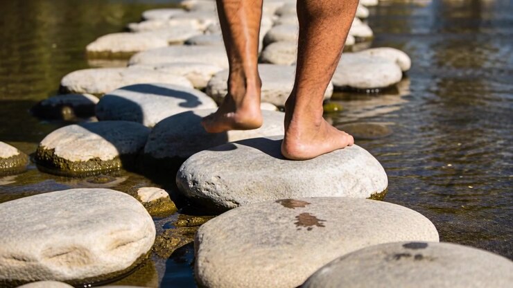 Uomo a piedi nudi appoggia il primo piede, attraversando un ruscello su pietre piatte, dirigendosi verso la riva opposta.