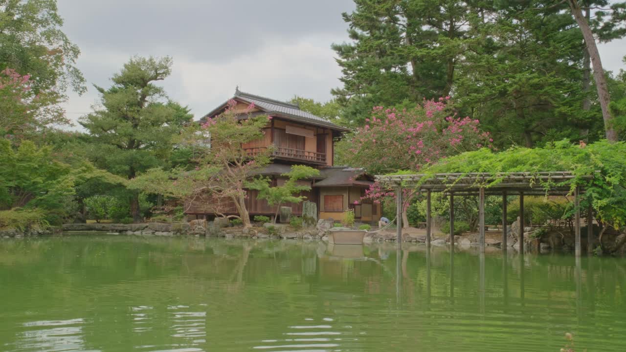 Wide shot of a traditional Japanese wooden villa next to a tranquil pond, with lush garden, blooming trees, and a shaded wooden pergola. Serene and timeless atmosphere.