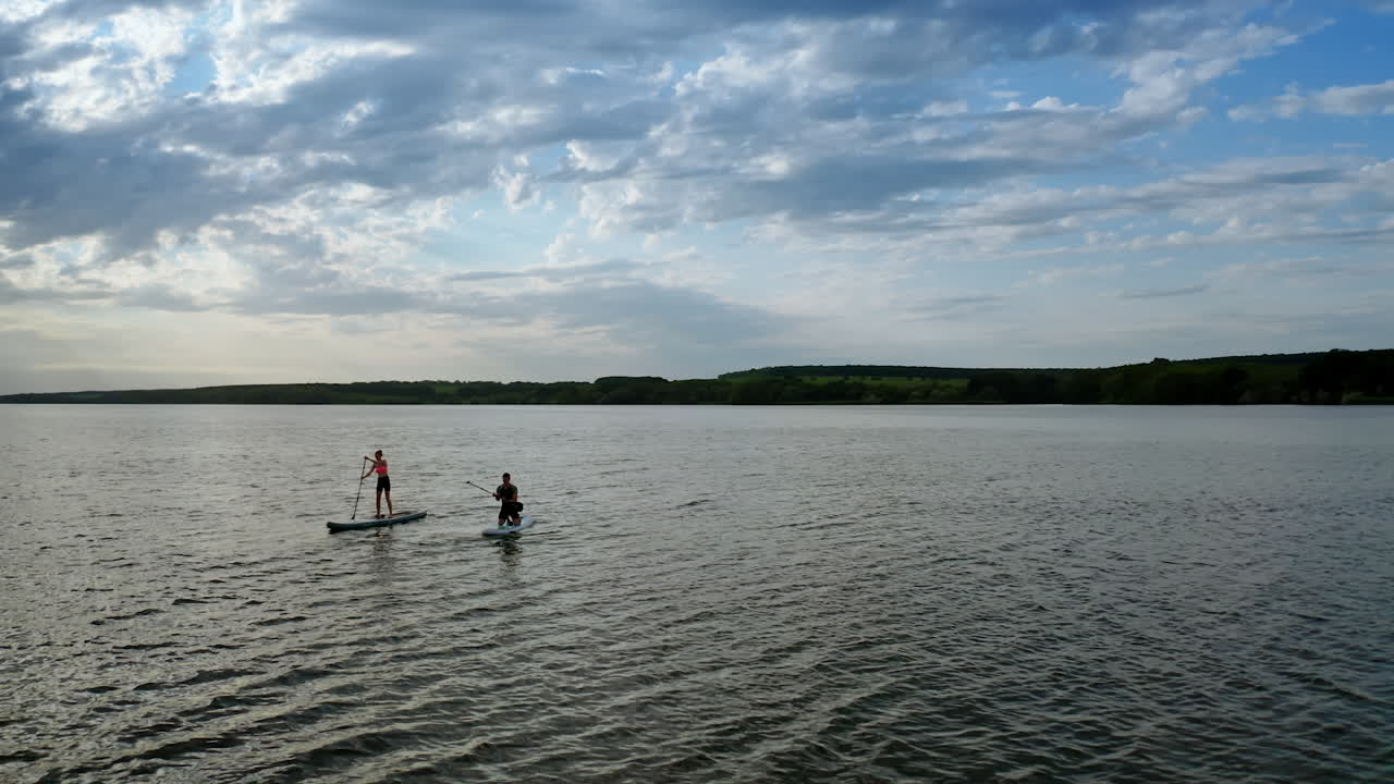 People spend time on water in the evening. Young man and woman sailing on river on boards with oar in summer at dusk. Orbital view.