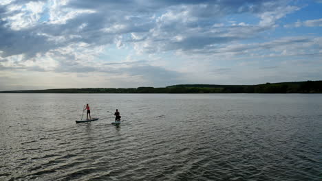 people spend time on water in the evening. young man and woman sailing on river on boards with oar in summer at dusk. orbital view.