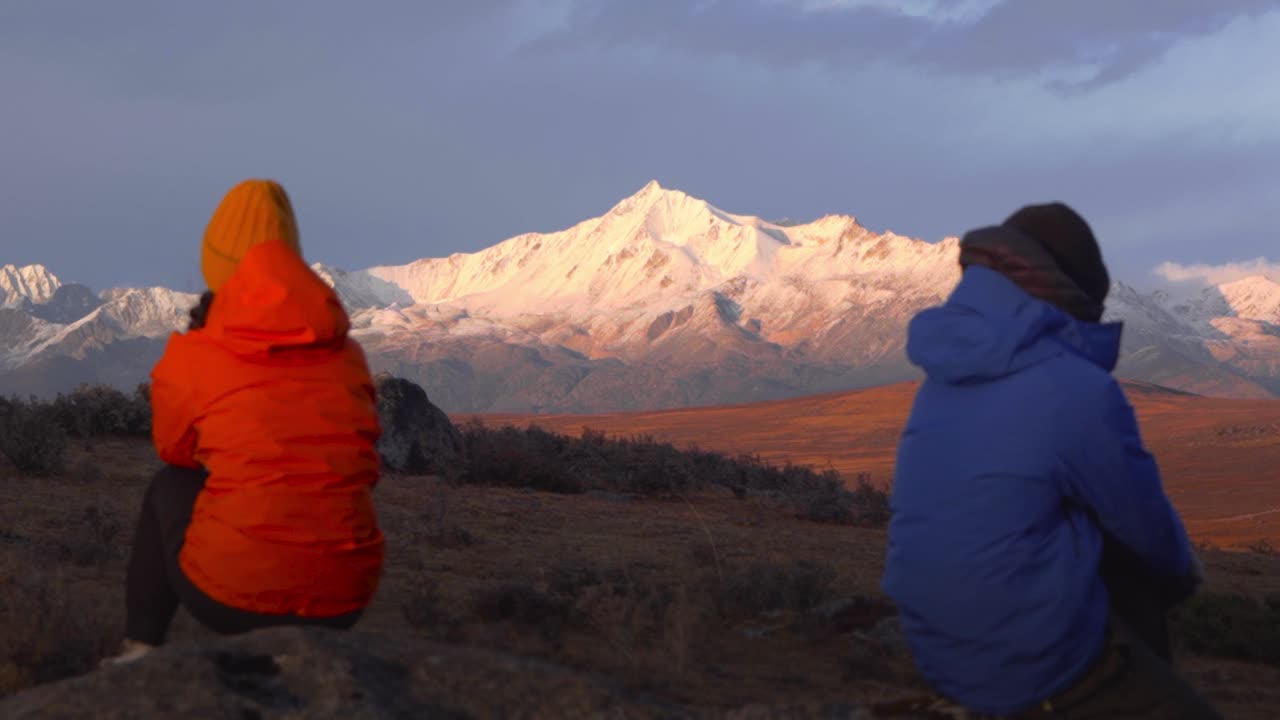 dos personas con chaquetas naranjas y azules se sientan mirándose el uno al otro mientras el resplandor alpino se establece en la montaña nevada de sichuan yala