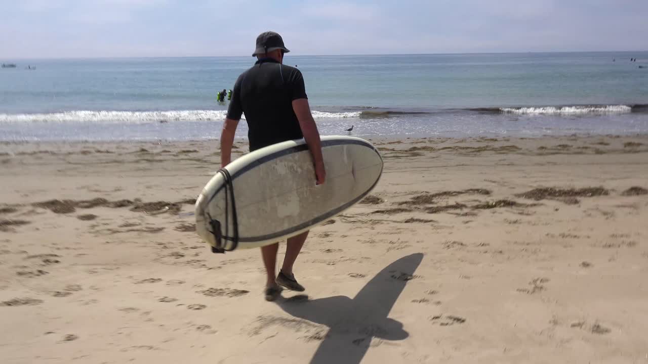 un surfista de mediana edad lleva su tabla por la playa y al agua en el sur de california