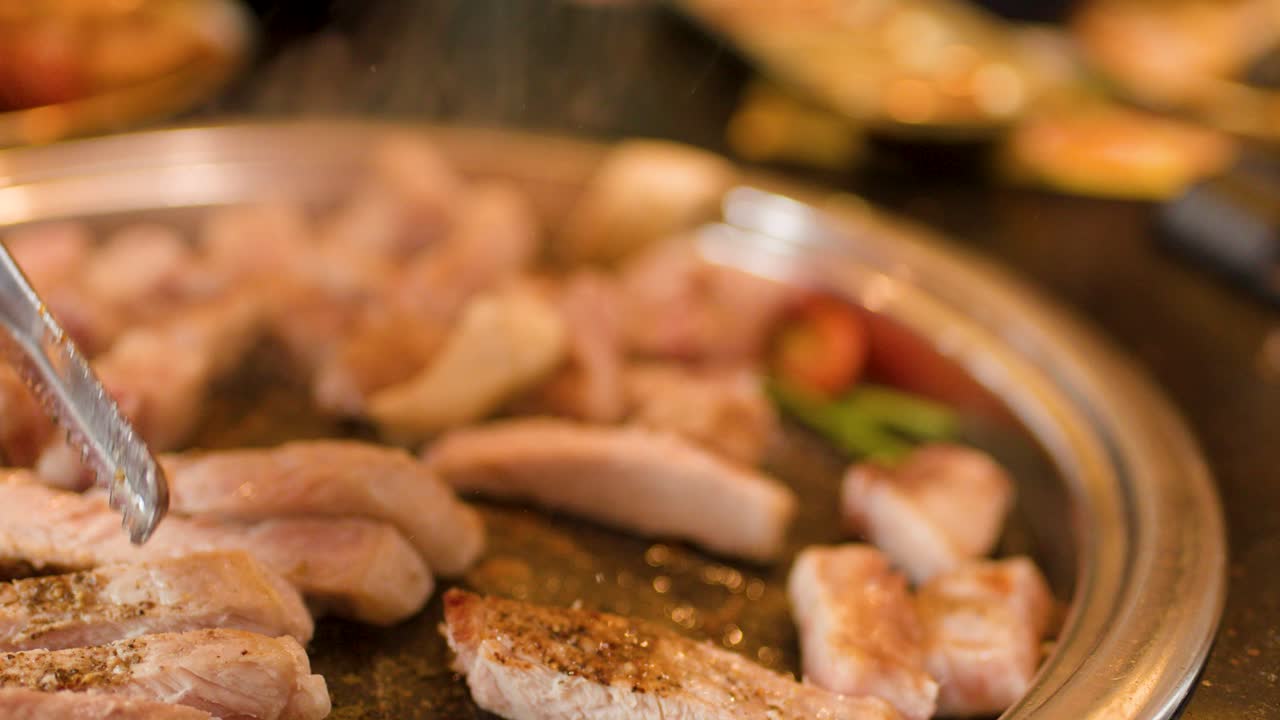 Sliced pork belly grilled on a hot plate, tongs turning meat, steam rising, warm lighting
