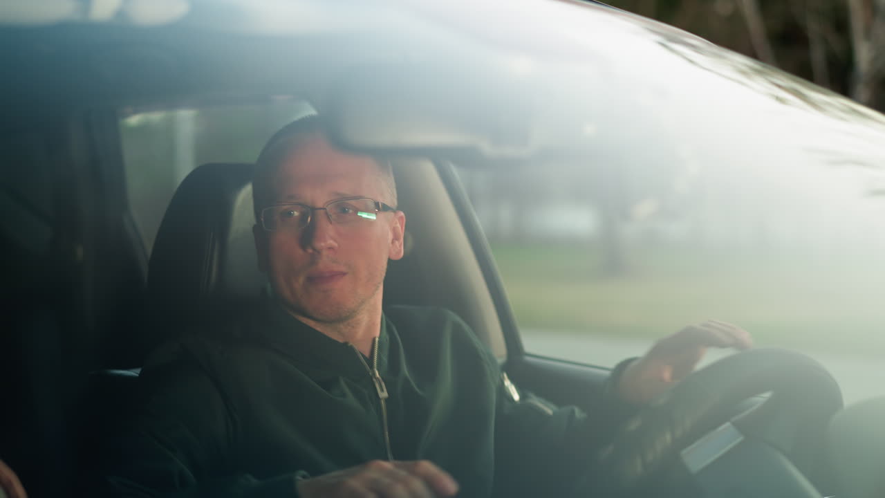 A man is seated in the driver's seat of a car, wearing a green jacket and glasses, gently dancing to music