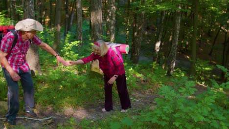 Senior-woman-on-hikinig-adventure-trip-holding-mans-hand-helping-her-climb-on-mountain-hill-in-wood