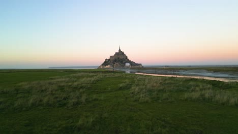 Luftdrohnenaufnahme-Des-Mont-Saint-Michel,-Der-Bei-Wunderschönem-Orangefarbenen-Sonnenuntergang-In-4k-über-Gras-Aufsteigt