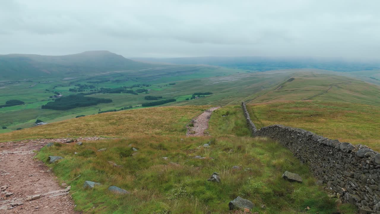 A winding hiking trail alongside a dry stone wall on a green hillside, overlooking a vast valley and distant mountains under an overcast sky