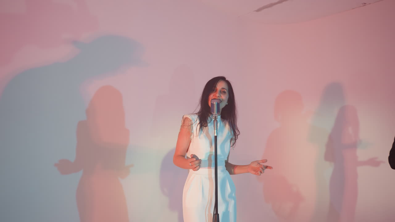 A vocalist wearing a white gown sings passionately while moving her hands. The camera zooms out, capturing her performance against a colorful background with dynamic lighting and shadows