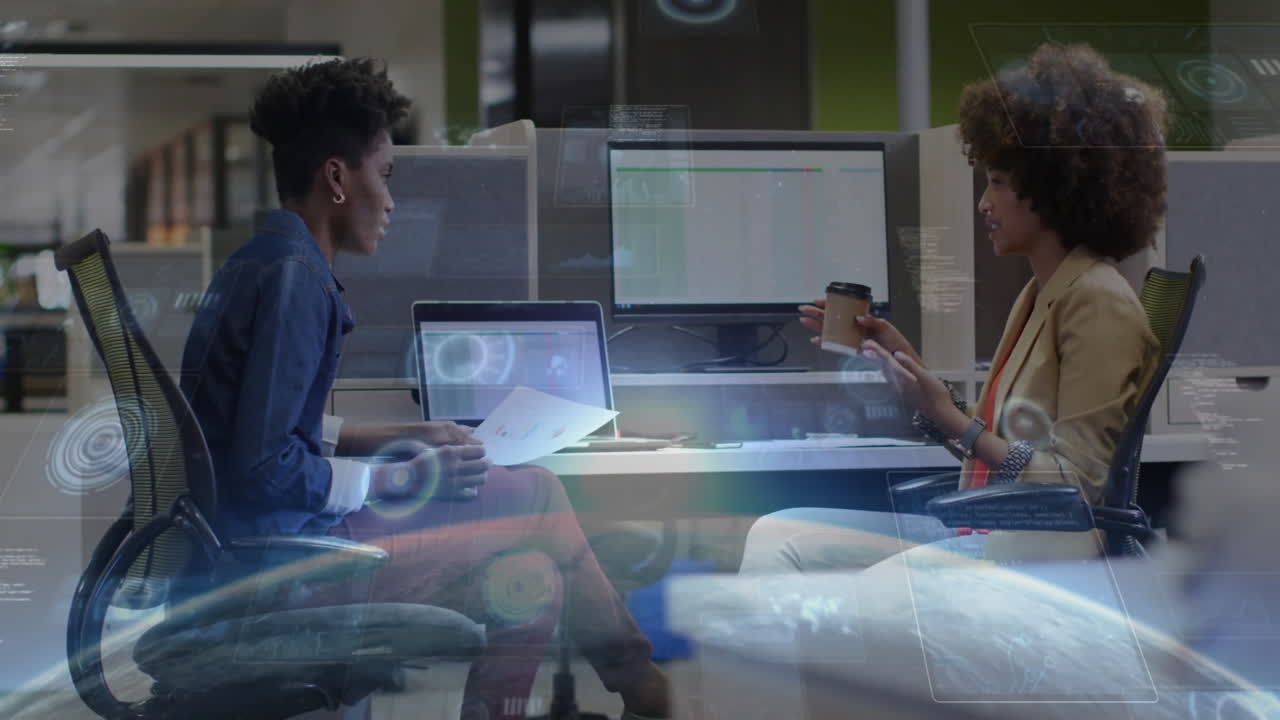 Two women reviewing charts on laptop at modern business office desk, showing floating data graphs