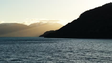 Silhouette-of-sloping-hills-down-to-water-surface-with-beams-of-light-cast-on-water